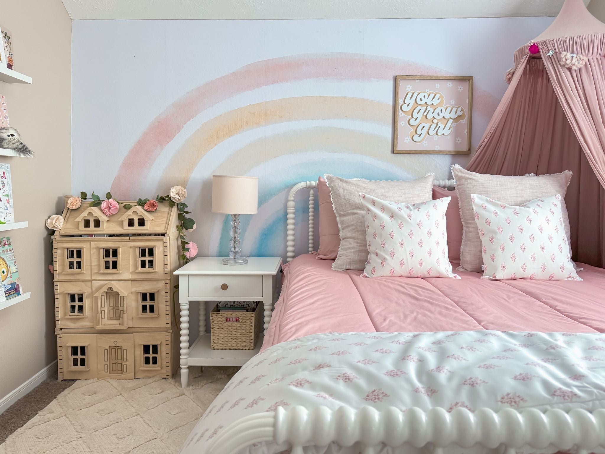 
      A cozy, pastel-themed girl's bedroom featuring pink Beddys bedding, patterned pillows, a nightstand, a wooden dollhouse adorned with pink flowers, and a whimsical rainbow mural on the wall.
    