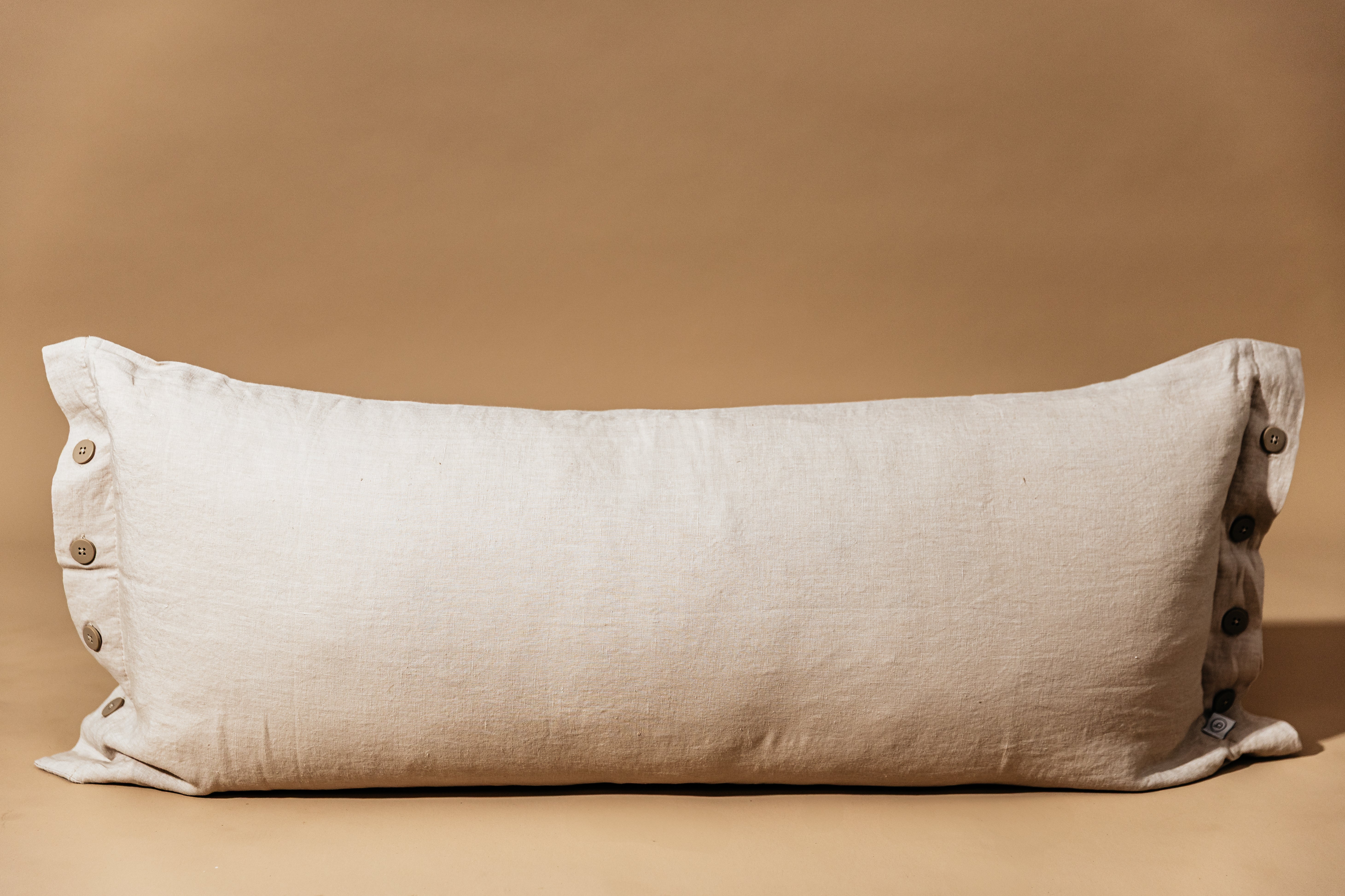 A lumbar linen pillow with buttons on each side sitting on a brown backdrop.