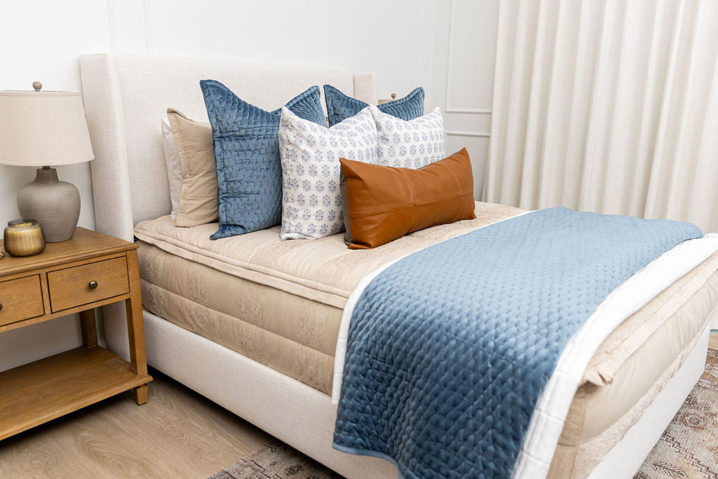 A queen bed made with tan zipper bedding and an assortment of blue and leather bedding accessories