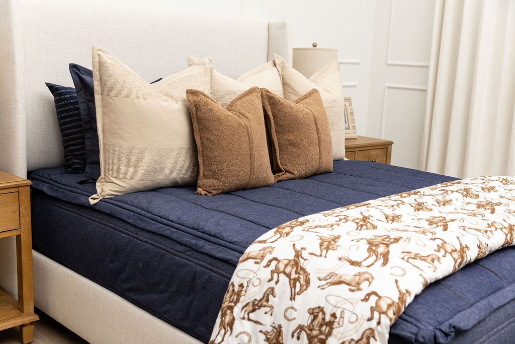 A brightly lit bedroom with a bed made with dark denim blue zipper bedding and an assortment of neutral pillows and a cowboy western printed blanket.