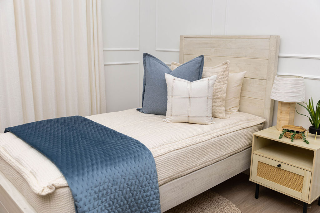 A bed made with cream waffle textured zipper bedding and an assortment of neutral green and pink throw pillows and blanket.