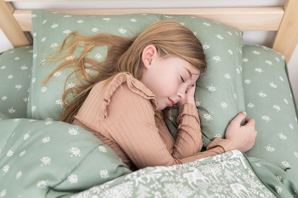 A young girl in tan pajamas sleeping in a bed made with sage green woodland creatures and trees kids zipper bedding.