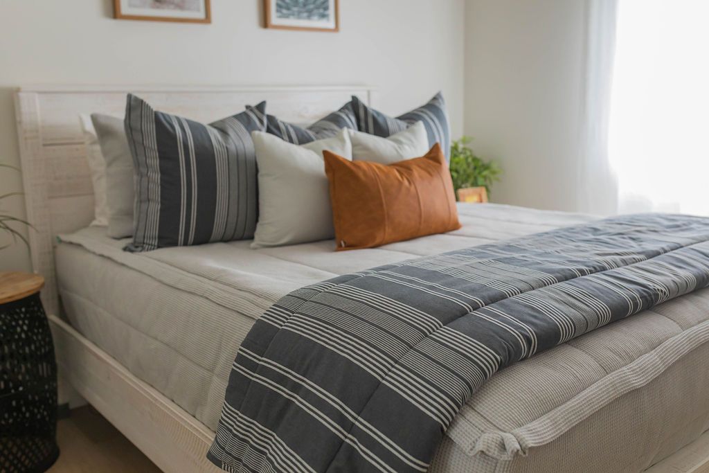 A brightly lit bedroom with a bed made with light gray zipper bedding and an assortment of neutral pillows and blanket.