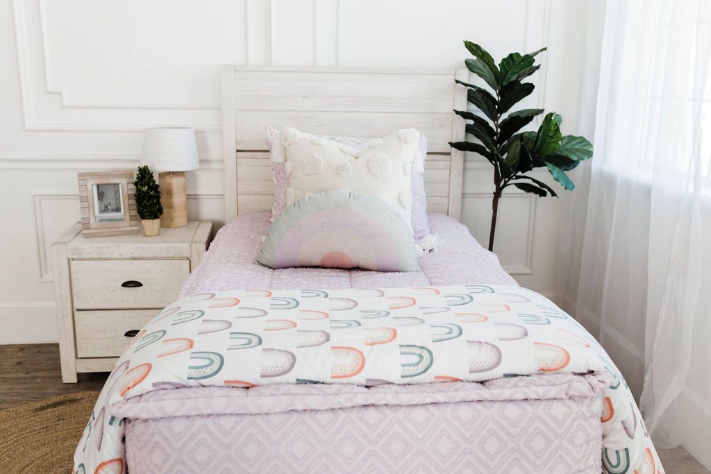 A brightly lit bedroom with a bed made with purple textured zipper bedding and a cream and pastel rainbow plush pillow as well as a rainbow pattern blanket.