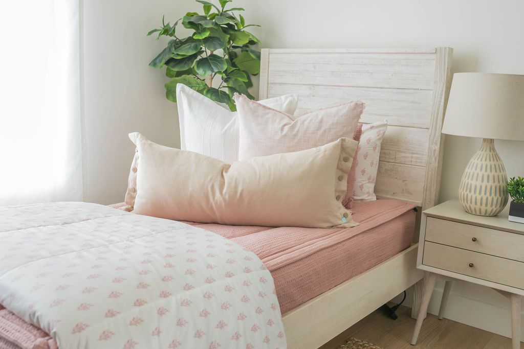 A bed made with woven pink zipper bedding and an assortment of neutral and pink pattern throw pillows and blankets.