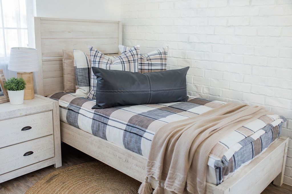 A bed made with cream, charcoal, and tan plaid zipper bedding and an assortment of neutral and leather throw pillows and blankets.