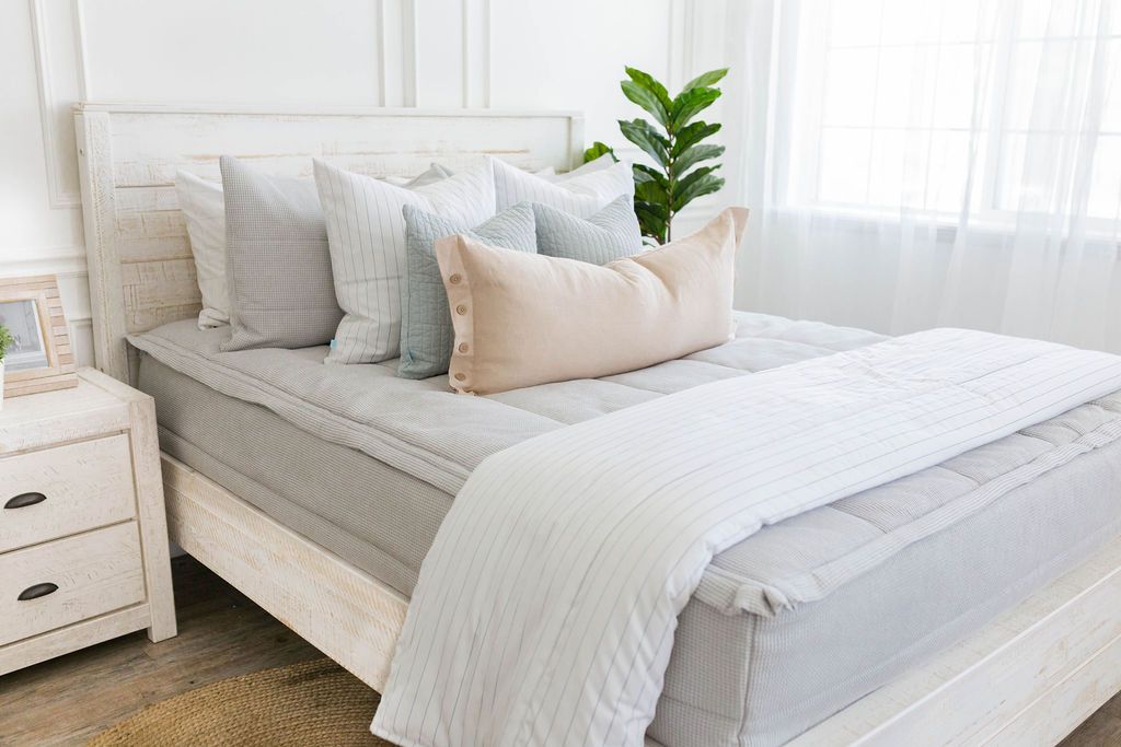 A lightly lit bedroom with a bed made with light gray waffle texture zipper bedding and an assortment of neutral and striped pillows and blanket.