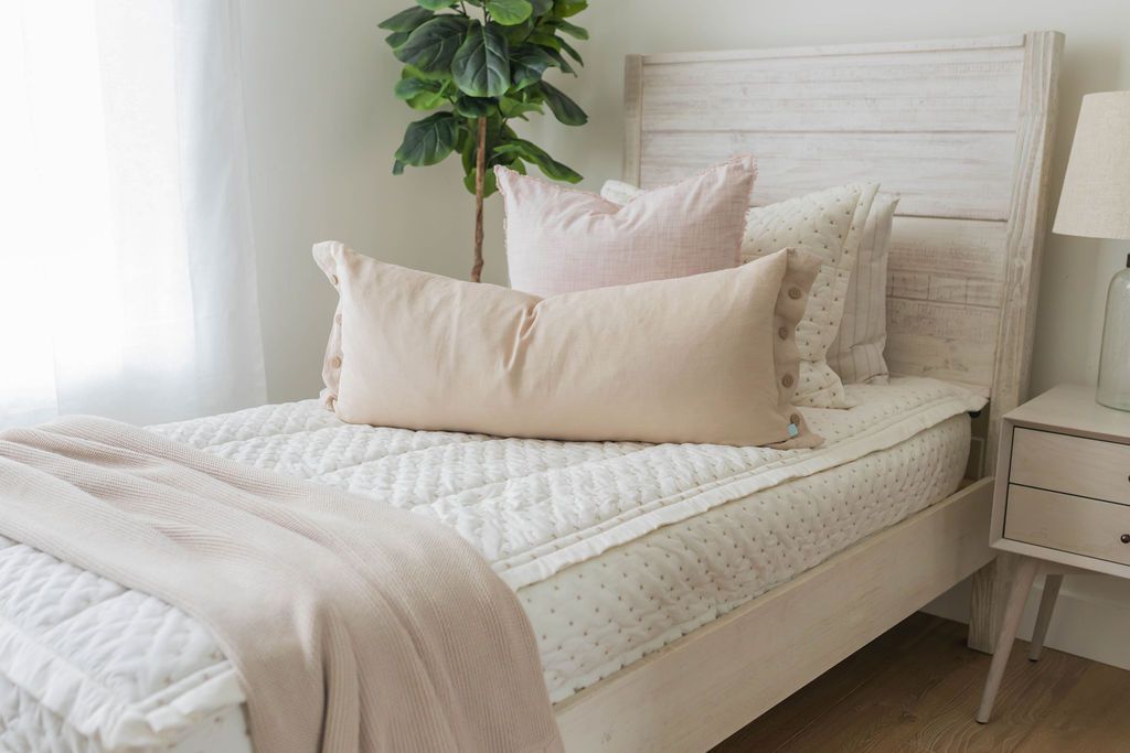 A lightly lit bedroom with a bed made with cream embroidered zipper bedding, decorated with neutral throw pillows and blankets.
