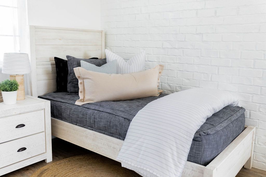A bed made with dark charcoal gray zipper bedding and an assortment of neutral and plaid throw pillows and blanket.