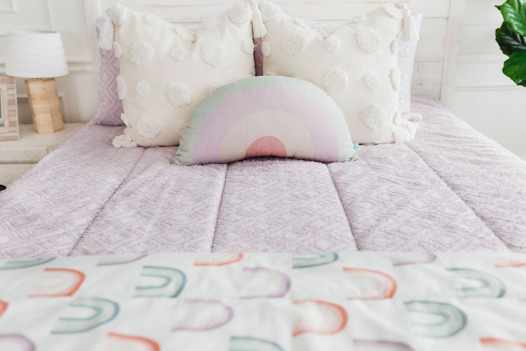 A close up view of a bed made with purple textured zipper bedding and neutral and rainbow pattern pillows and matching blanket.