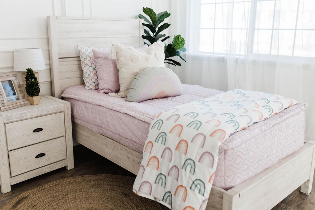 A brightly lit bedroom with a bed made with purple textured zipper bedding and an assortment of neutral and rainbow pattern pillows and blanket.