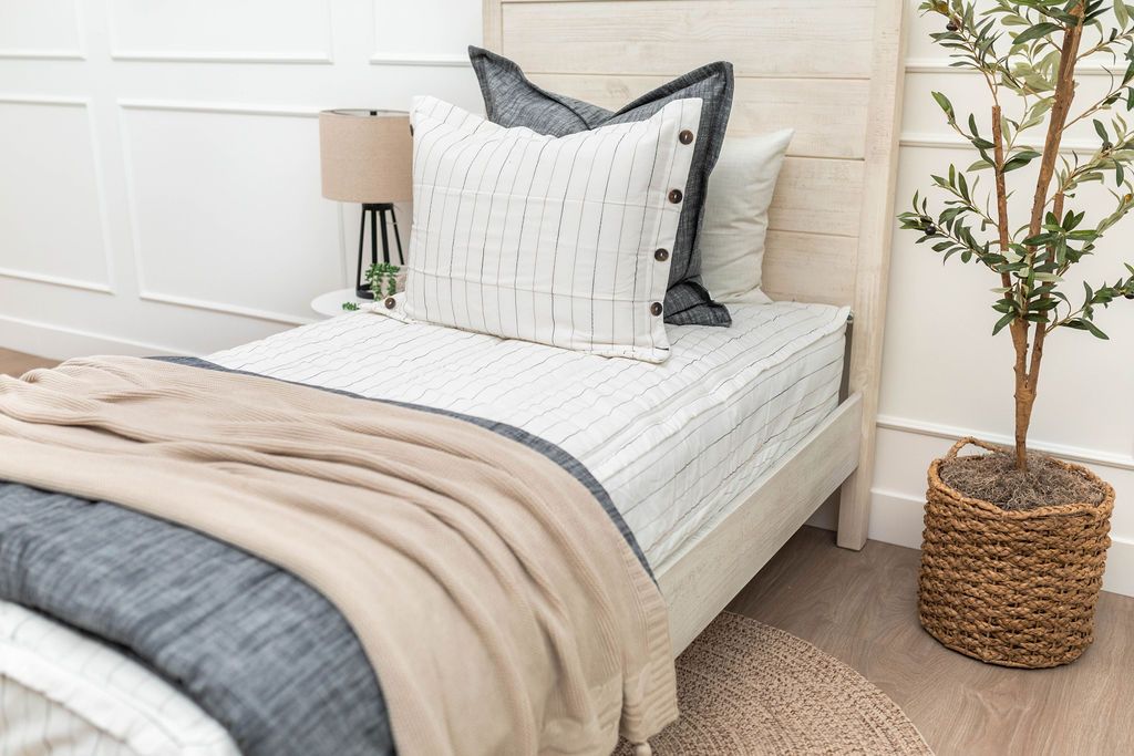 A bed made with white and pinstripes zipper bedding and an assortment of neutral and dark gray pillows and blanket.